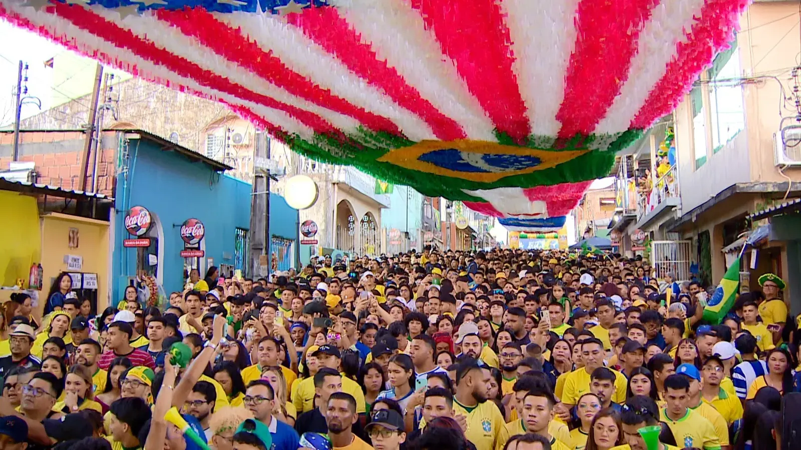 Multidão de torcedores reunidos na Rua da Copa em Manaus, com decorações temáticas e um telão transmitindo a partida da Seleção Brasileira.