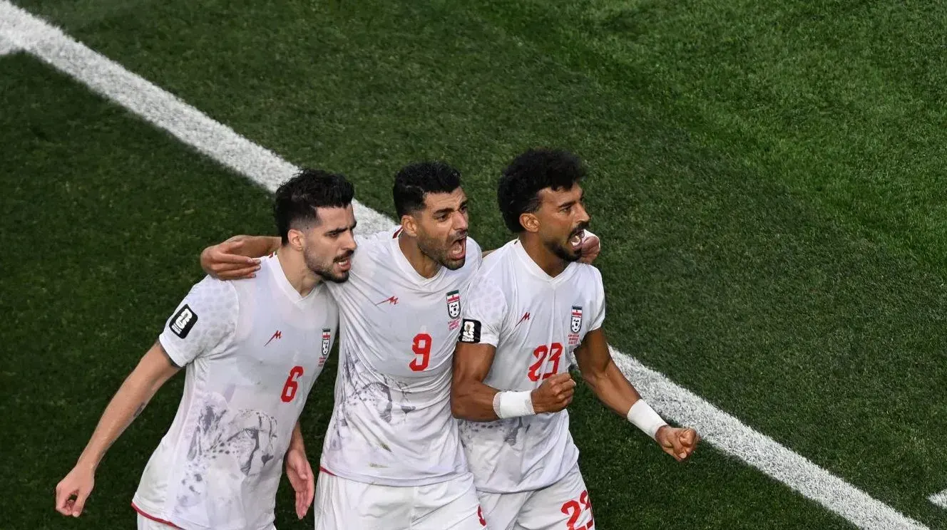 Jogadores de futebol do Irã e da Nova Zelândia disputando a bola em campo.