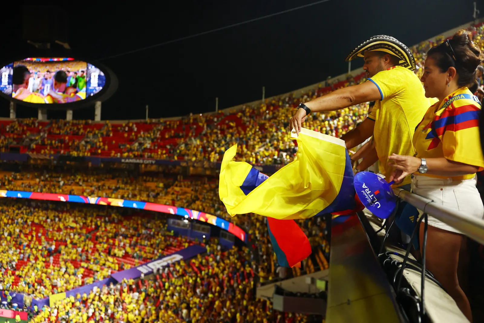 Torcedores colombianos comemoram e vibram em um estádio de futebol, vestindo a camisa amarela da seleção e ostentando bandeiras do país.