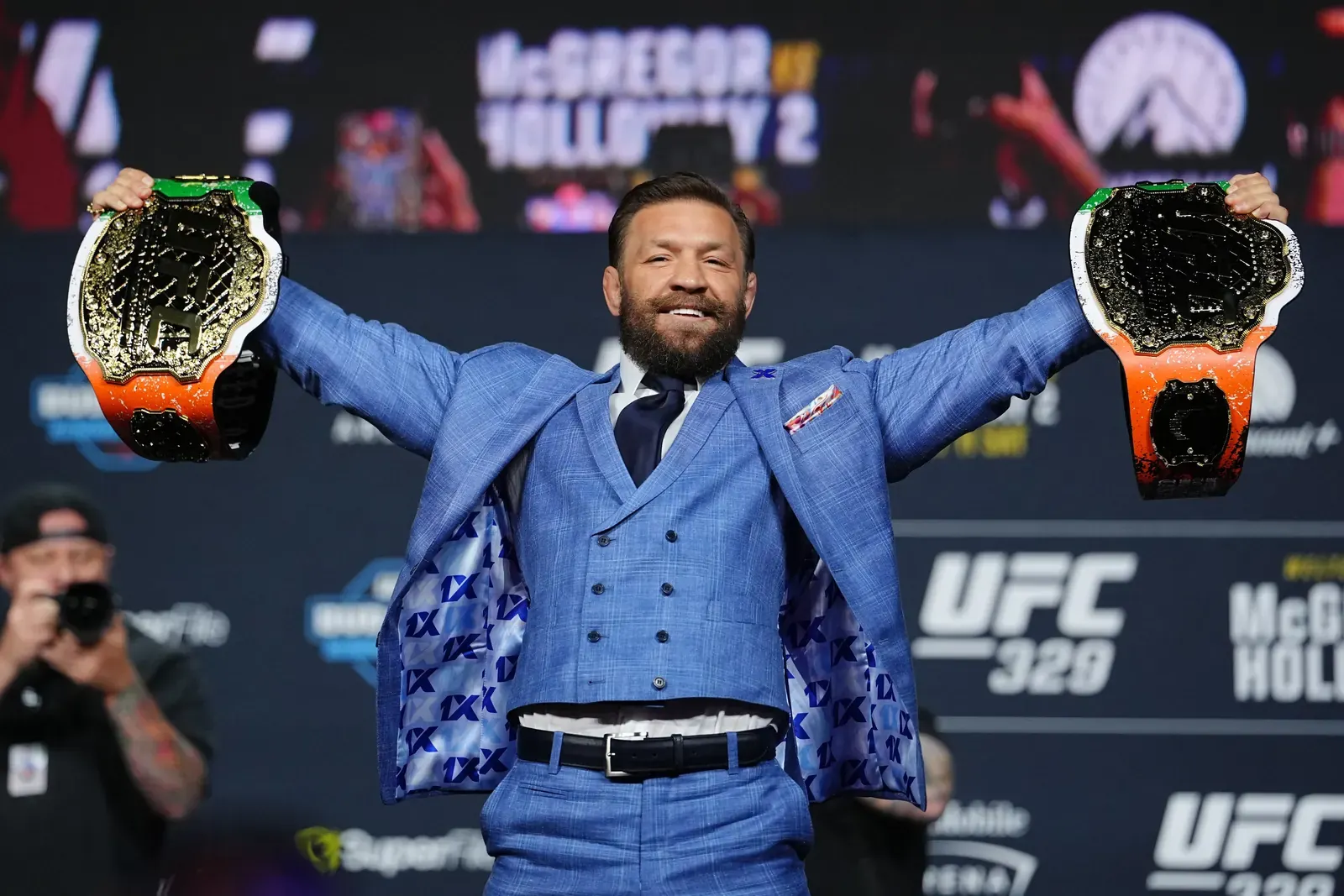 Conor McGregor sorrindo durante coletiva de imprensa do UFC, com cinturões ao fundo.